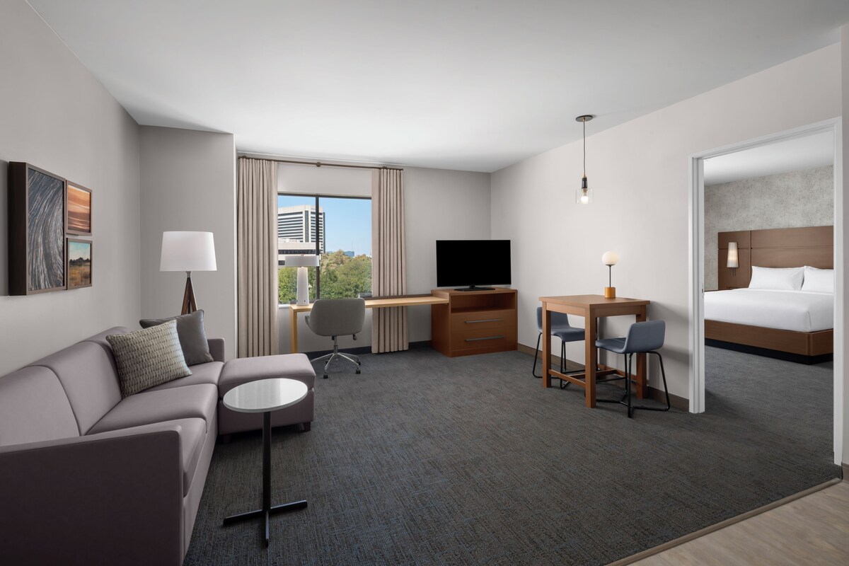 A hotel room with a sofa, a desk, a TV, a bed, and a view of the city.