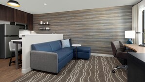 A modern hotel room with a sofa, a small table, a desk, a kitchen area, and a patterned carpet.