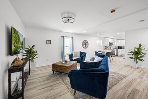 Ein modernes Wohnzimmer mit einem blauen Sofa, einem Couchtisch und einem Fernseher auf einem Schrank. Im Hintergrund befindet sich eine Küche mit weißen Schränken und einem Essbereich.