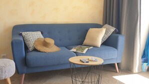 A blue sofa with patterned cushions, a round wooden side table with books, and a fluffy stool. - Charming apartment with a panoramic view of the sea. 5 minutes from the beaches (Valle-di-Campoloro)