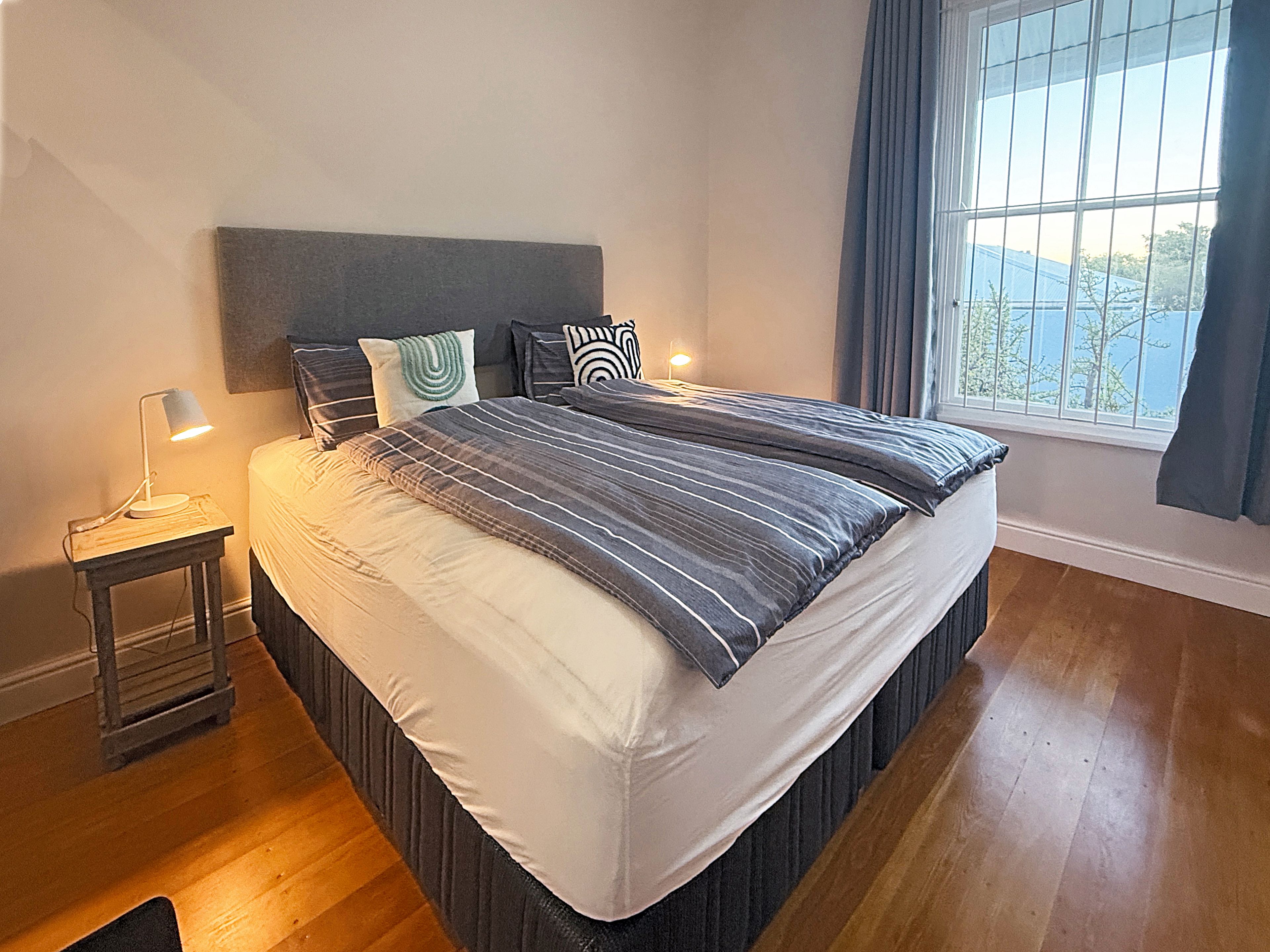 A neatly made bed with a headboard, a bedside table with a lamp, and a window with curtains.