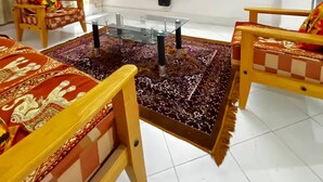 A living room with a flat-screen TV, two wooden chairs, a glass coffee table, a patterned rug, and a ceiling fan.