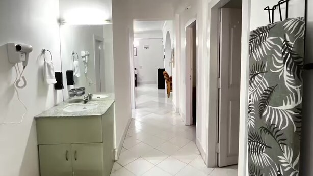 A hallway with a white tiled floor, a mirror, a sink, and a patterned wall hanging.