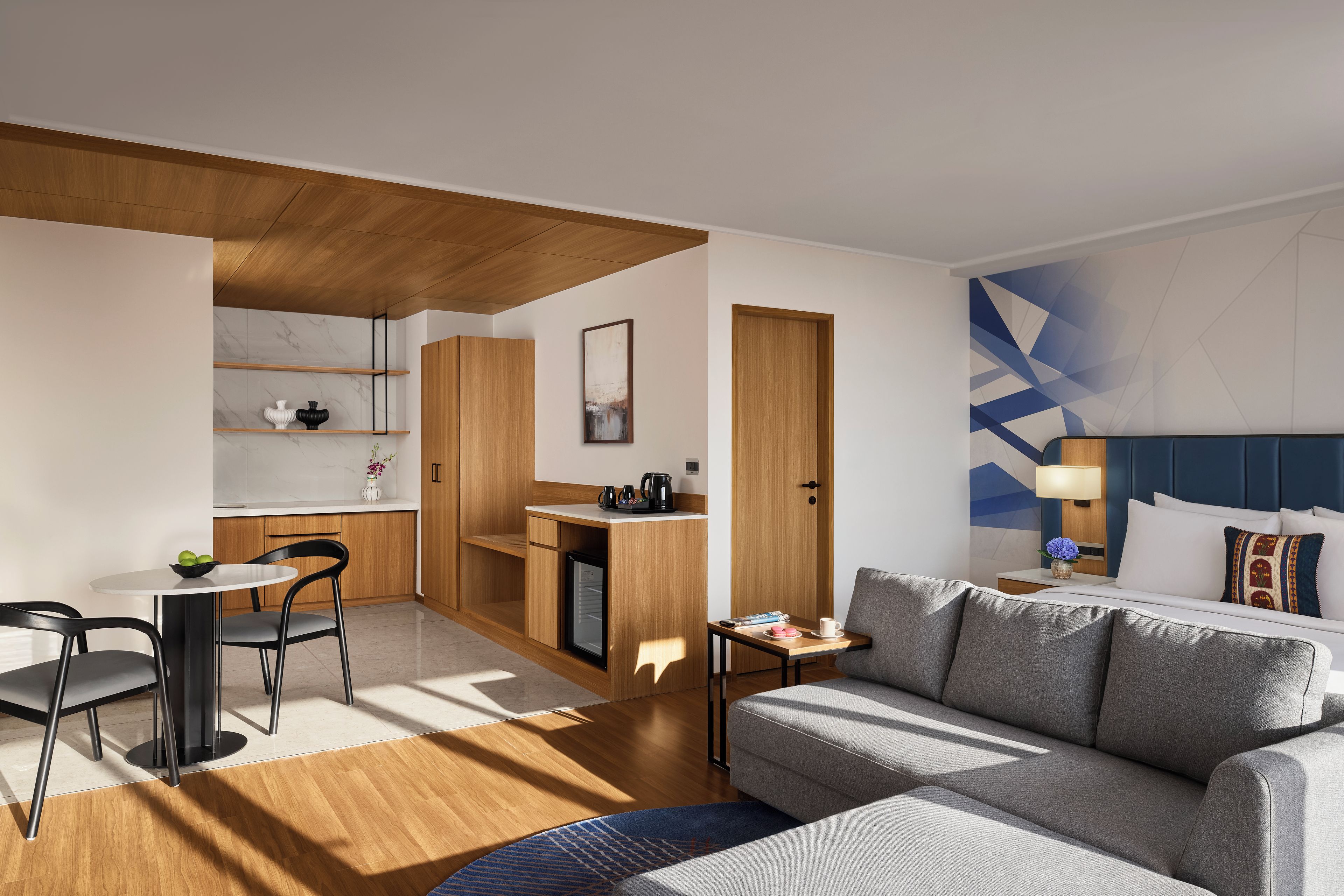 Quarto de hotel moderno com sofá, mesa de jantar com cadeiras, cozinha compacta e cama com cabeceira.