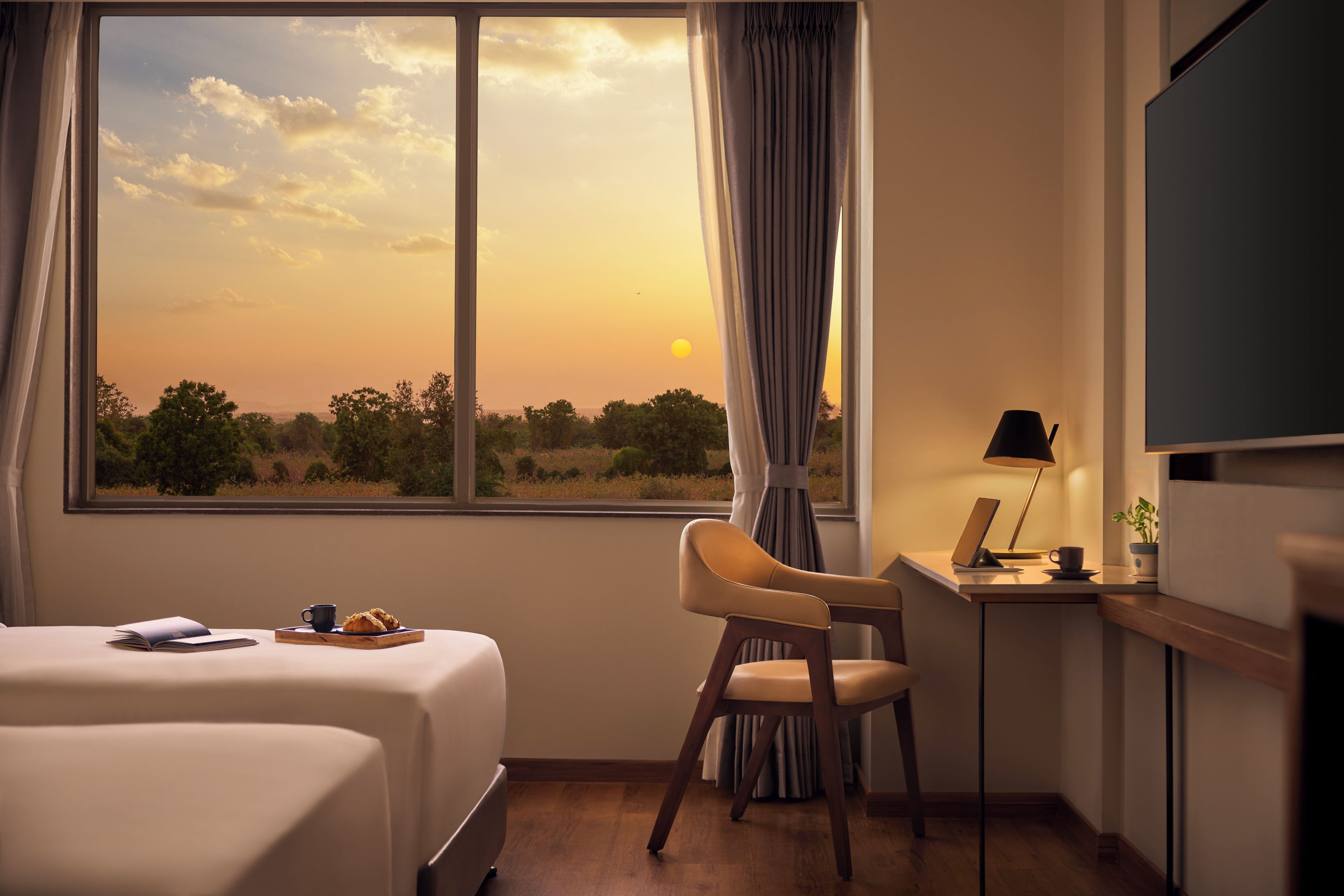 A hotel room with a large window, a bed, a desk with a laptop, a chair, and a television.