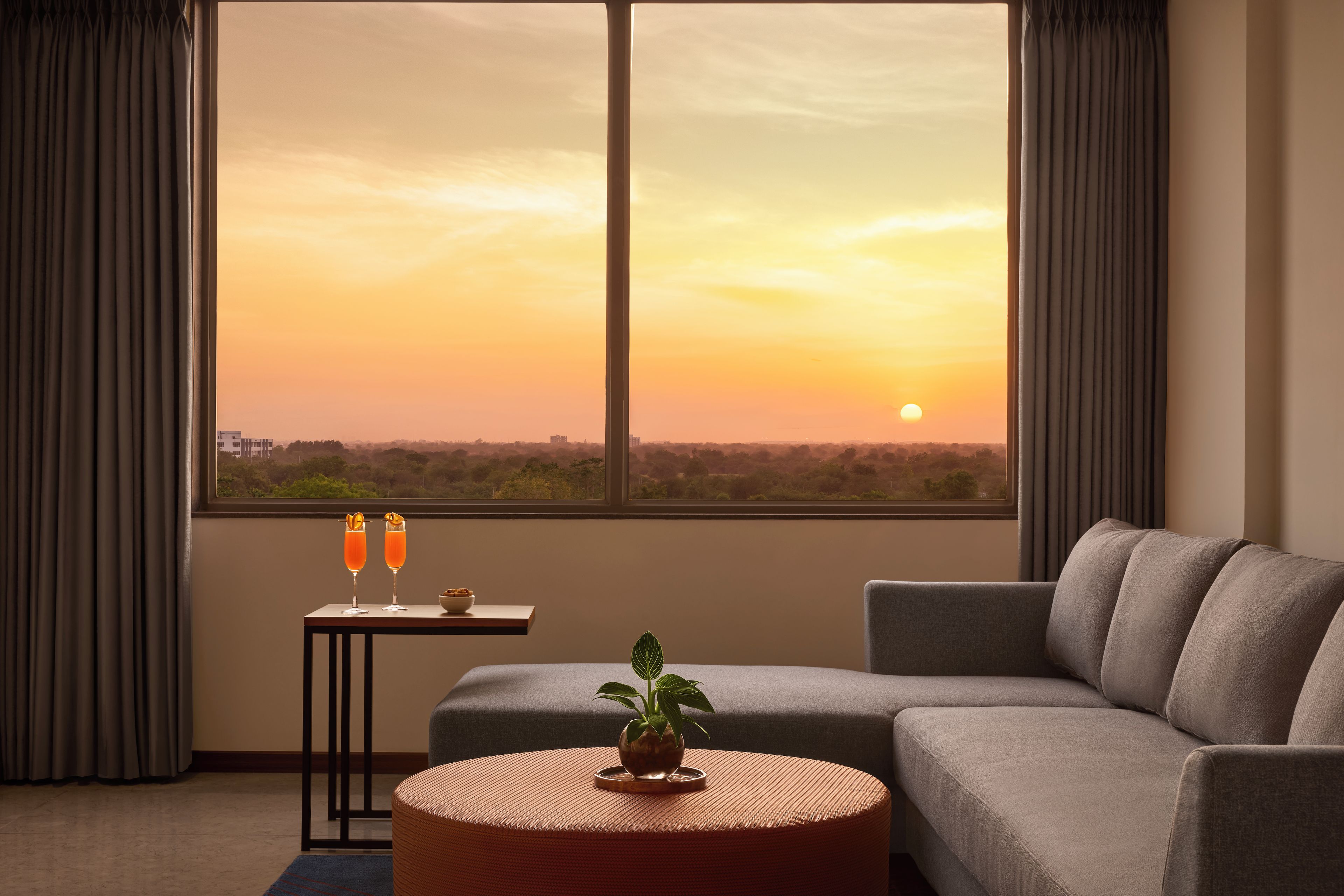 A modern living room with a large window, a grey sofa, a small round table with a plant, and two glasses of orange beverage.