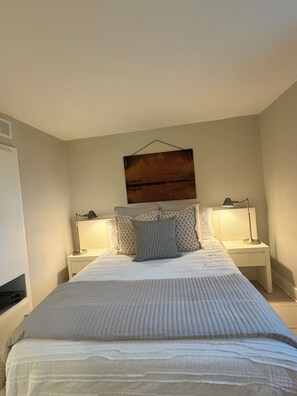 Cama bem arrumada com edredom branco e travesseiros, duas luminárias de cabeceira e uma obra de arte na parede acima da cama.