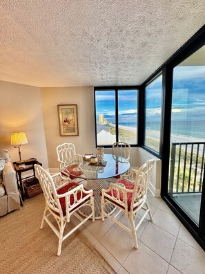 Quarto com mesa de vidro, cadeiras brancas com almofadas vermelhas, luminária, quadro emoldurado e vista para a praia e oceano através de grandes janelas.