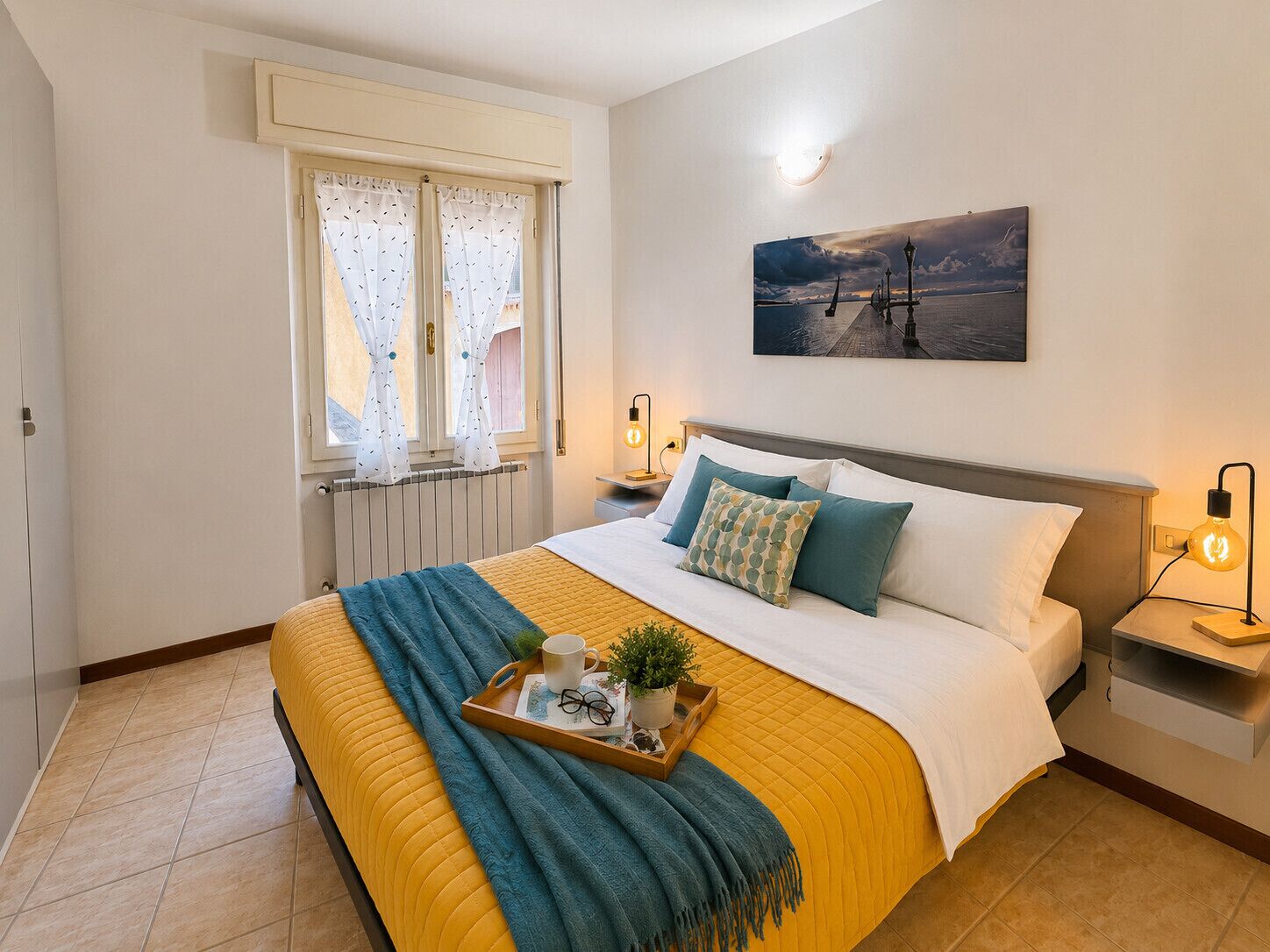 Ein Schlafzimmer mit Bett, Nachttischen, einem Fenster mit Vorhängen, einem Bild an der Wand und einem Tablett mit Gegenständen auf dem Bett.