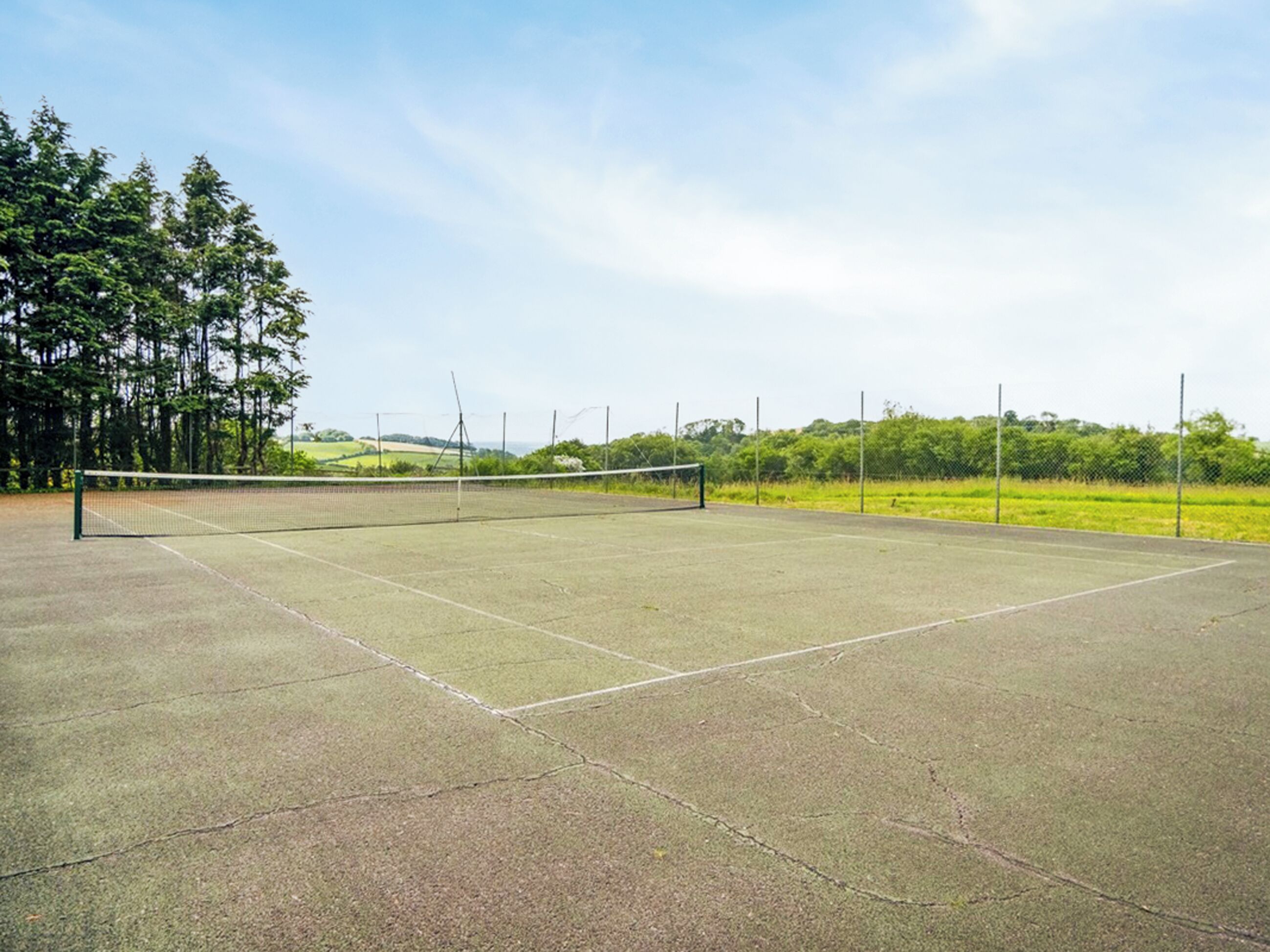 Cancha de tenis al aire libre con red, rodeada de árboles y campos abiertos.