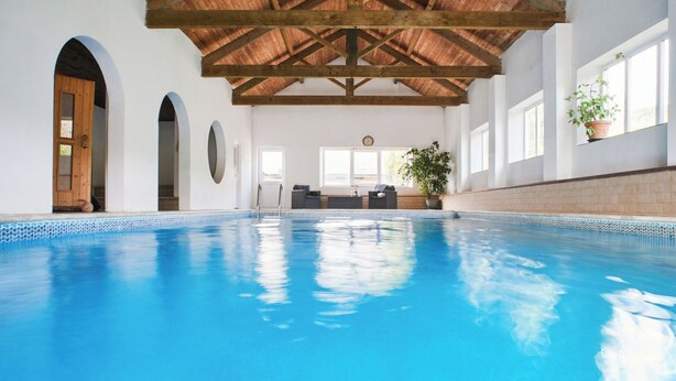 Una piscina interior con techo de madera, paredes blancas y una zona de estar con una planta en maceta.
