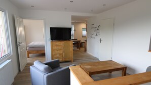 Un salon moderne avec un canapé, une table basse en bois, un téléviseur posé sur un meuble et un lit à l’arrière-plan.