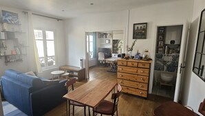 Sala de estar con un sofá azul, una mesa de comedor de madera y sillas, una zona de cocina con un armario de madera, y un baño con inodoro y lavamanos.