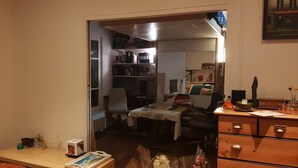 Una habitación con escritorio, silla y una pequeña mesa con mantel. Hay una estantería y un armario.