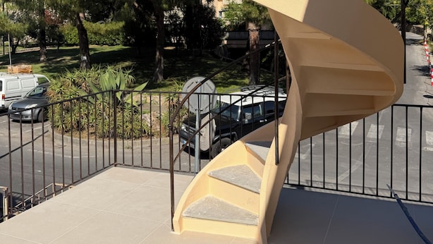 Una escalera en espiral con exterior color beige y barandillas de metal negro, que conduce a un nivel superior.