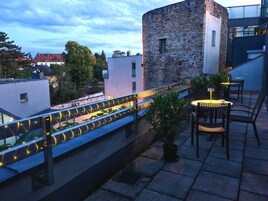 A rooftop terrace with a stone wall, a railing with string lights, and outdoor seating.