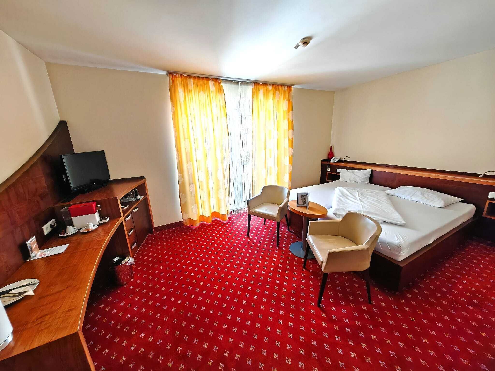 Een hotelkamer met een bed, twee stoelen, een bureau met een televisie en een roodgekleurd tapijt.