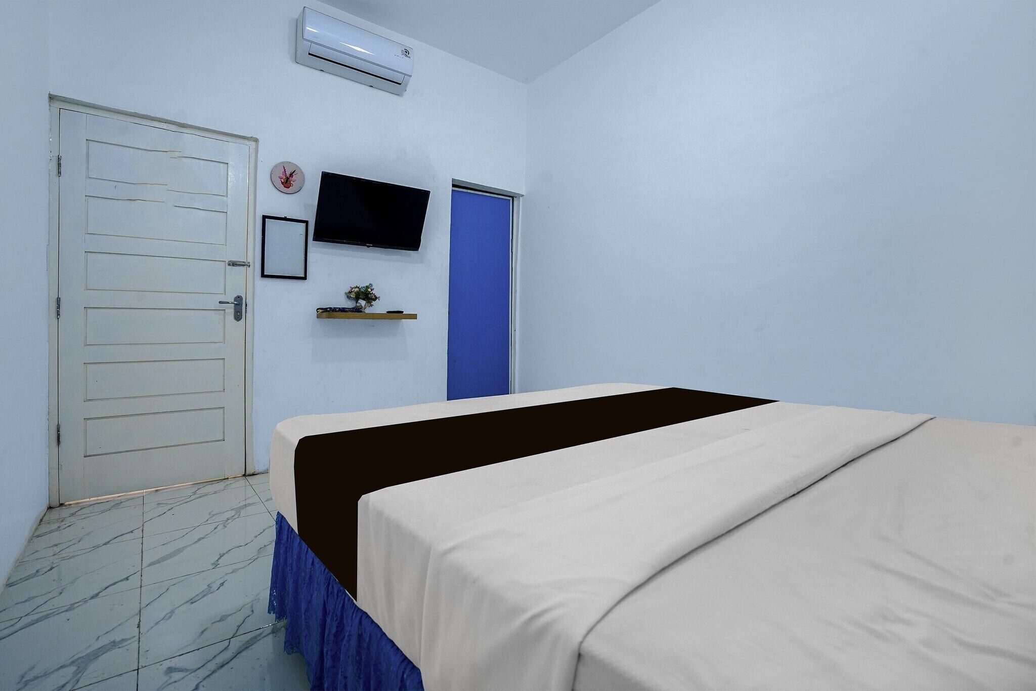 Una habitación con cama, puerta, televisor montado en la pared y un pequeño estante con una planta.
