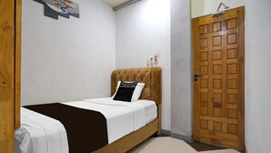 Un lit simple avec une tête de lit, une porte en bois et une petite étagère fixée au mur, sur laquelle sont posés quelques objets.
