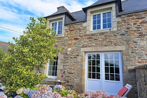A stone house with a large window and a door, surrounded by lush greenery and flowers. - Stone house in Brittany by the sea (Lannilis)