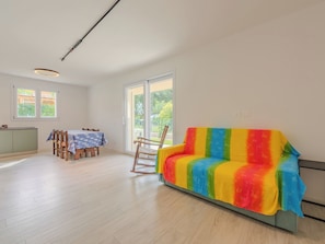 Ein geräumiger Raum mit Holzboden, einem bunten Sofa, einem Esstisch mit Stühlen und einem Fenster mit Blick auf Grünflächen.