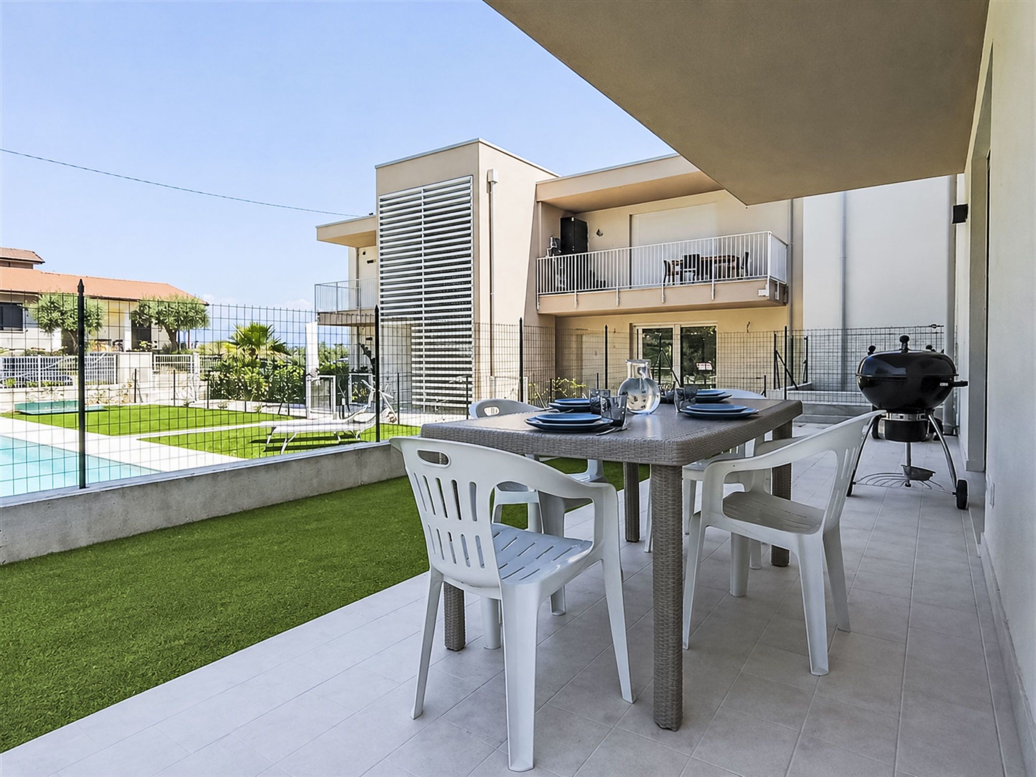 An outdoor patio with a table set for dining, a grill, and a view of a swimming pool.
