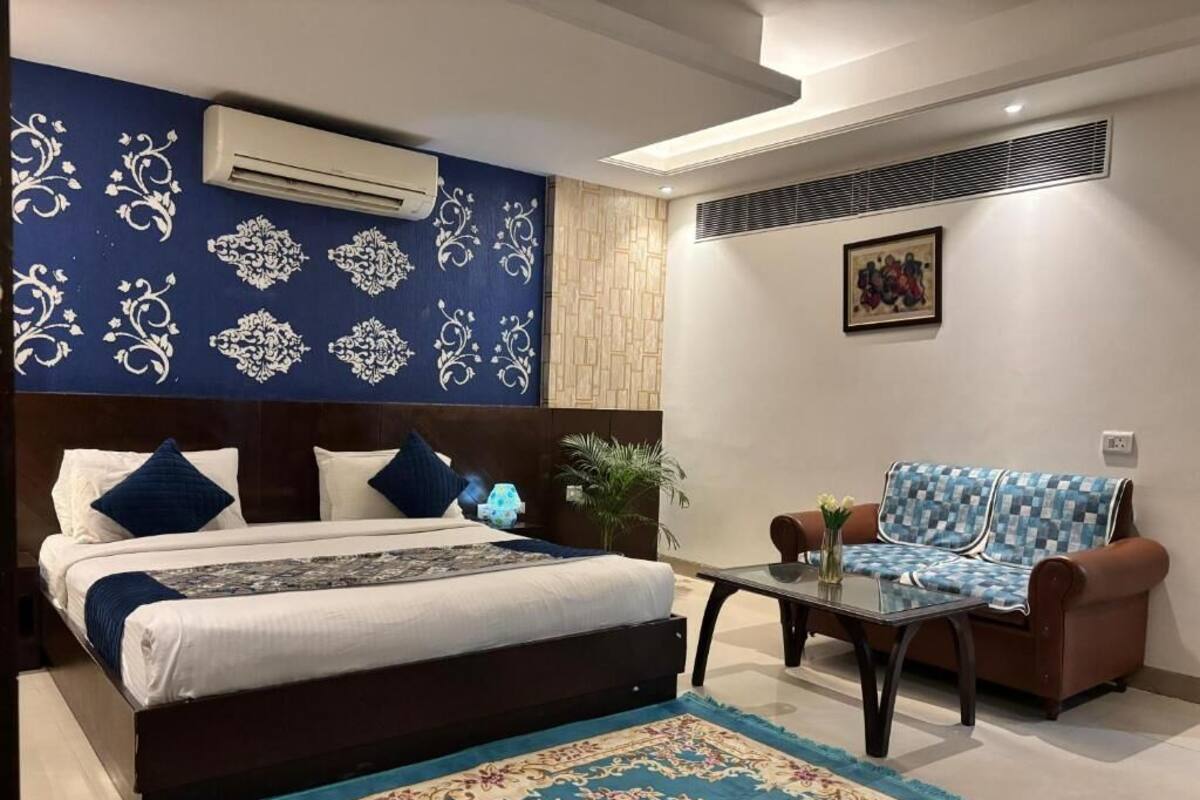 A bedroom with a bed, a sofa, a coffee table, an air conditioner, a wall-mounted artwork, and a patterned rug.
