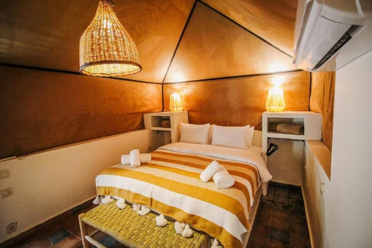 A cozy, compact room with a bed, two bedside tables, a hanging light fixture, and an air conditioning unit.