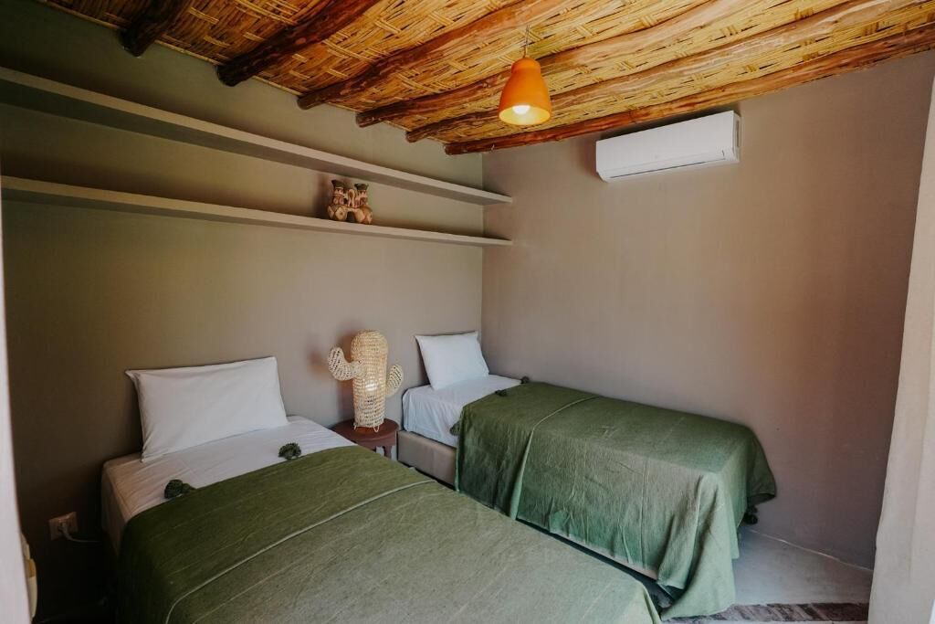 A room with two beds, a woven ceiling, a small shelf with decorative items, and a single hanging light fixture.