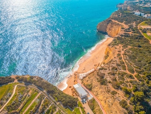 An aerial view of a coastal area with a beach, cliffside, and a building with a flat roof. - Villa Bloom Coast (Lagoa)