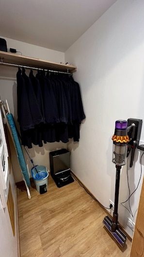 A small room with a wooden floor, a clothes rack with dark garments, a vacuum cleaner, and a blue mop bucket.