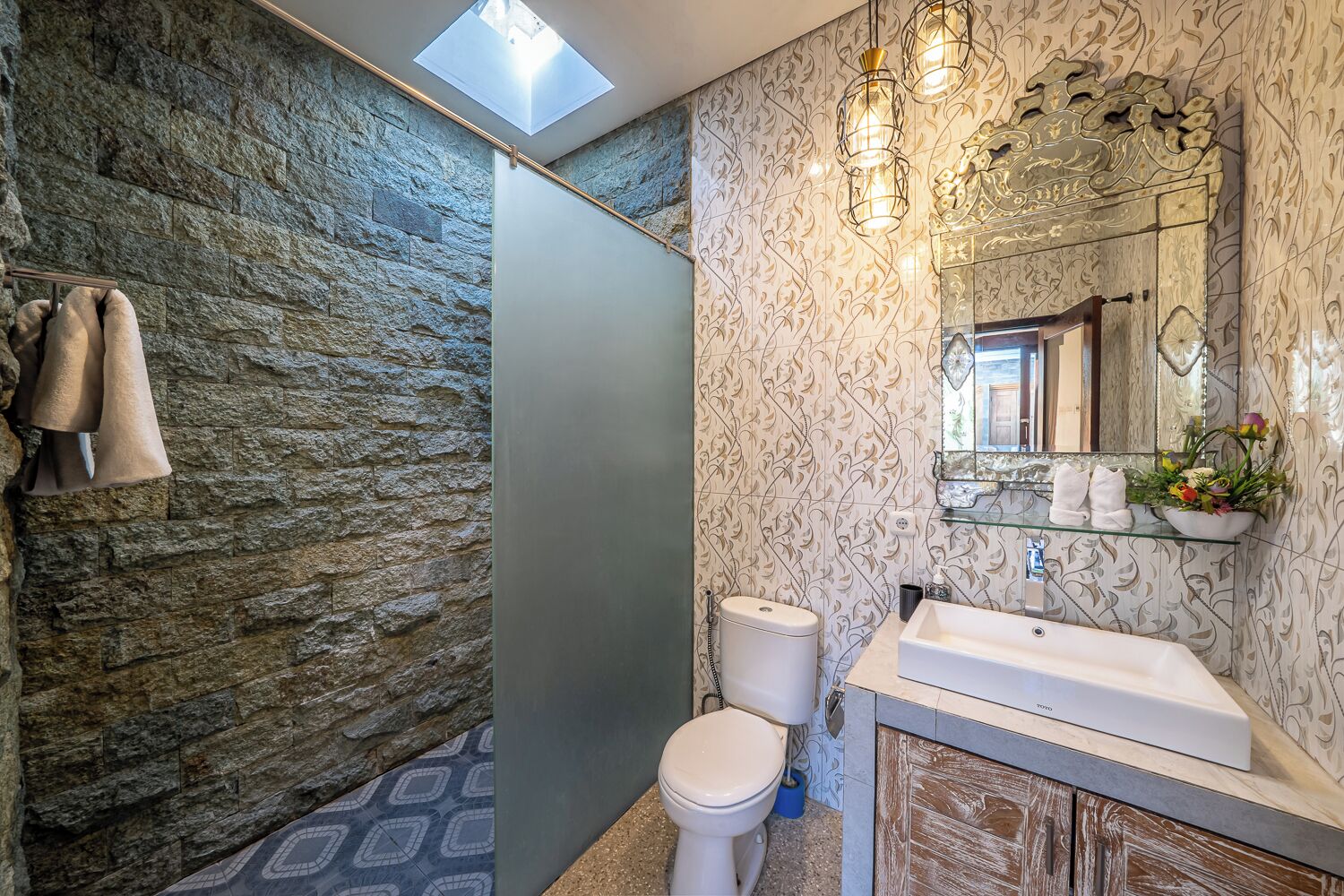 A bathroom with a stone wall, a skylight, a toilet, a sink, and a mirror with a decorative frame.