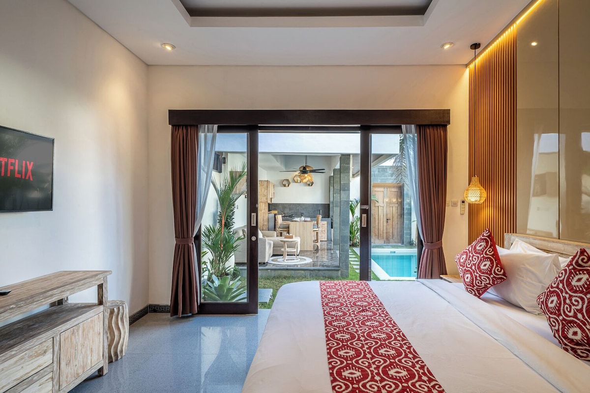 Ein Schlafzimmer mit einem großen Bett, einem Fernseher, einer hölzernen Bank und Blick auf einen Pool und eine Außenlounge.
