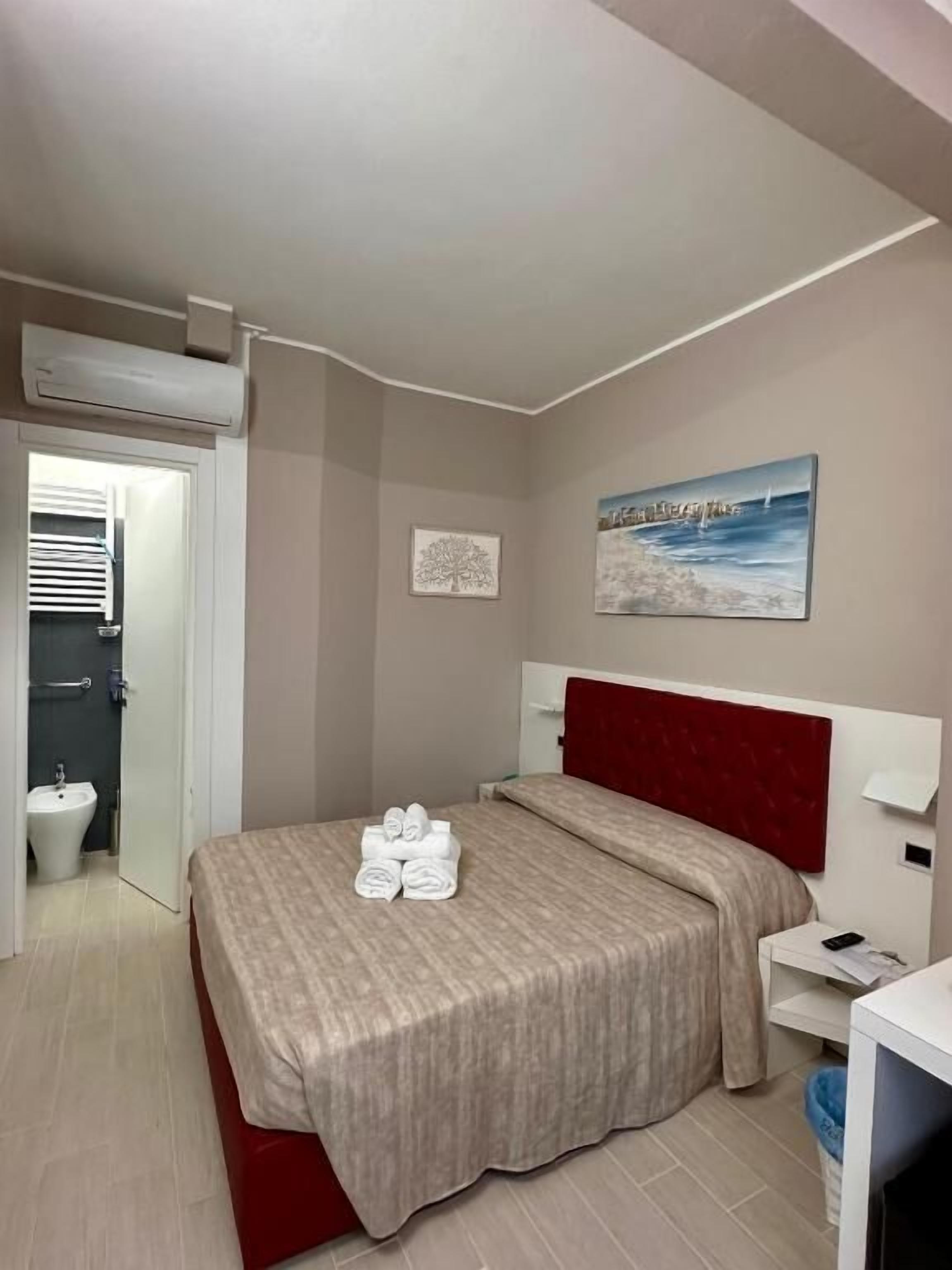 Habitación de hotel con cama, cabecera roja, un cuadro, mesita de noche y pared blanca.