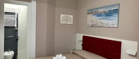 Habitación de hotel con cama, cabecera roja, un cuadro, mesita de noche y pared blanca.