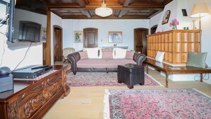 Ein Zimmer mit Holzbalkendecke, einem Kronleuchter, einem Bett mit gemusterten Betttextilien, einem Holzkasten, einem Fernseher und einem Schreibtisch mit Lampe.