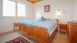 Ein Schlafzimmer mit einem hölzernen Bett, zwei Nachttischlampen, einem kleinen Nachttisch, einem Beistelltisch, einem dekorativen Kissen, einem gerahmten Bild an der Wand und einem Fenster mit Jalousien.