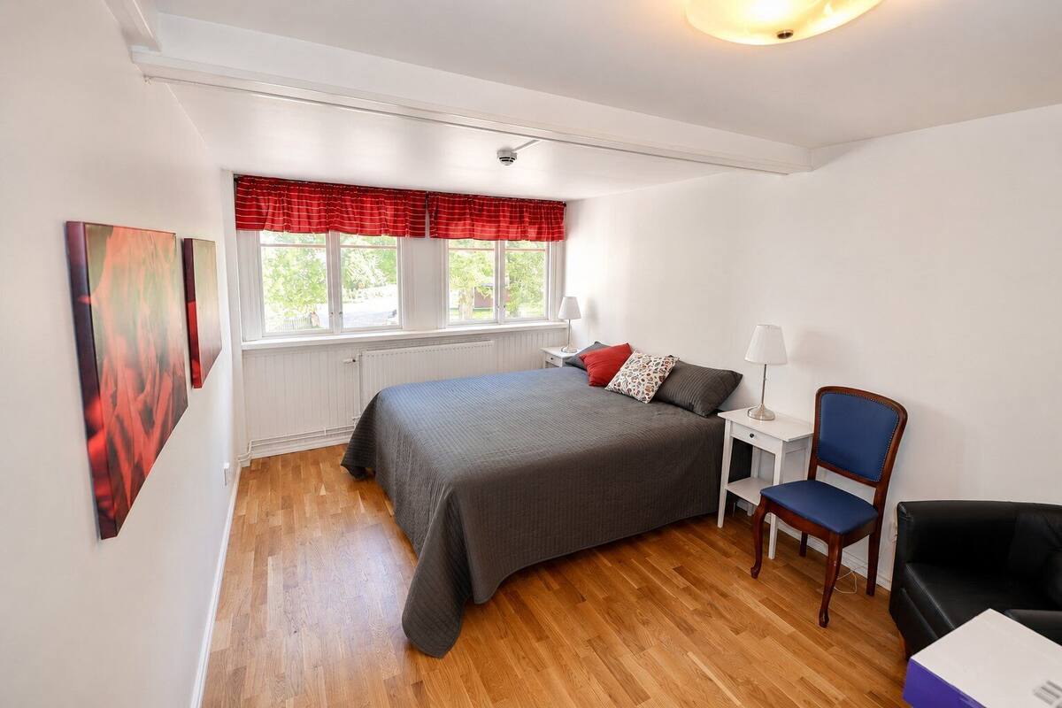 A bedroom with a bed, a chair, a nightstand, a window with curtains, and a wall with a painting.