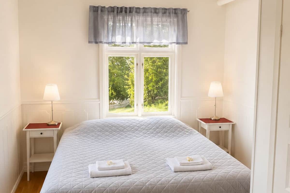 A neatly arranged bedroom with a large bed, two bedside tables with lamps, a window with a view of greenery, and a door on the right.