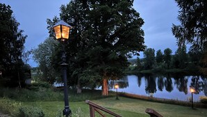 A wooden deck with a metal railing, a lamp post, and a calm lake reflecting trees. - Husby Wardshus (Dala-Husby)