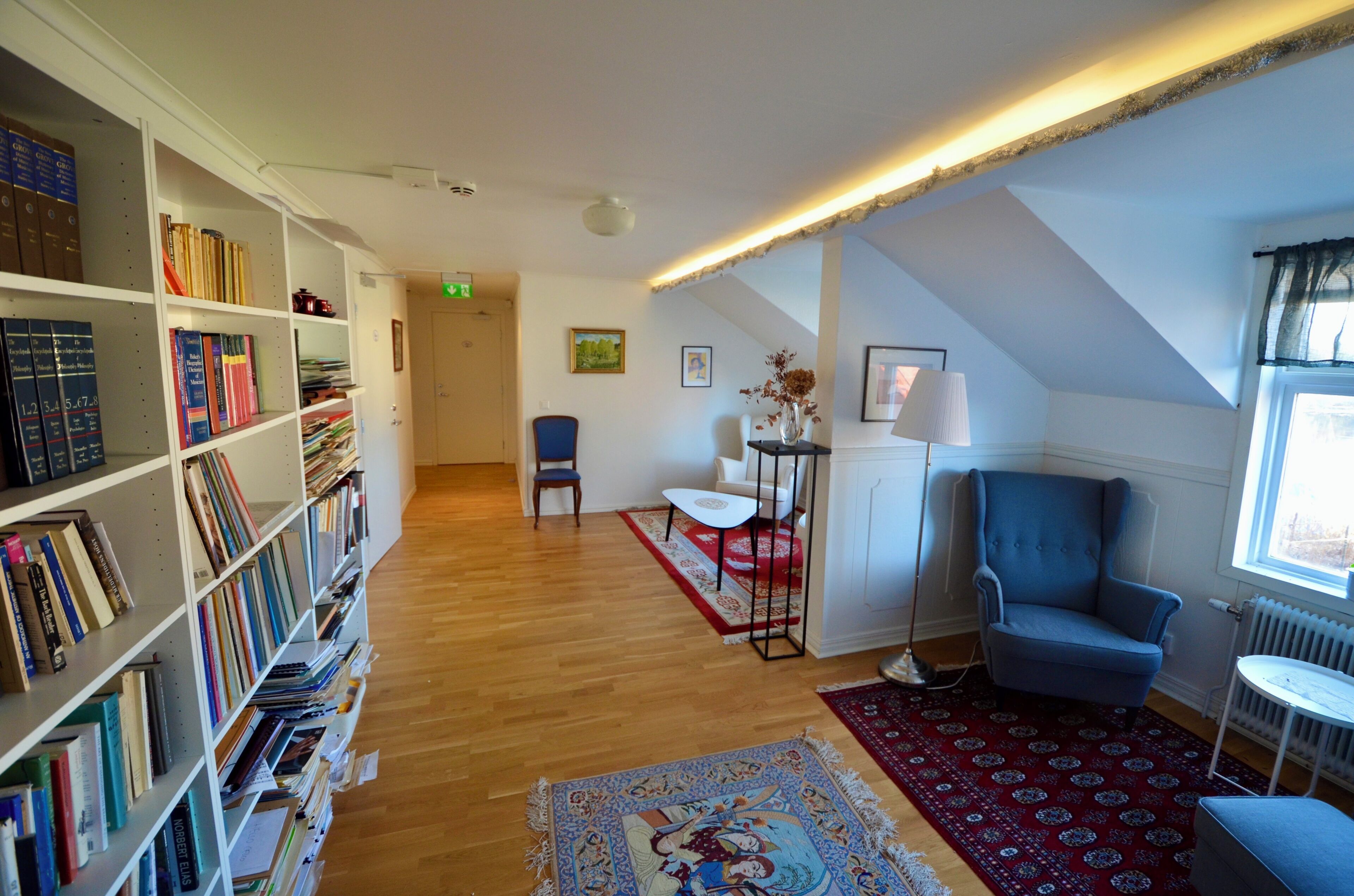Una habitación con una estantería, un sillón azul, una mesa blanca y un tapete rojo estampado.
