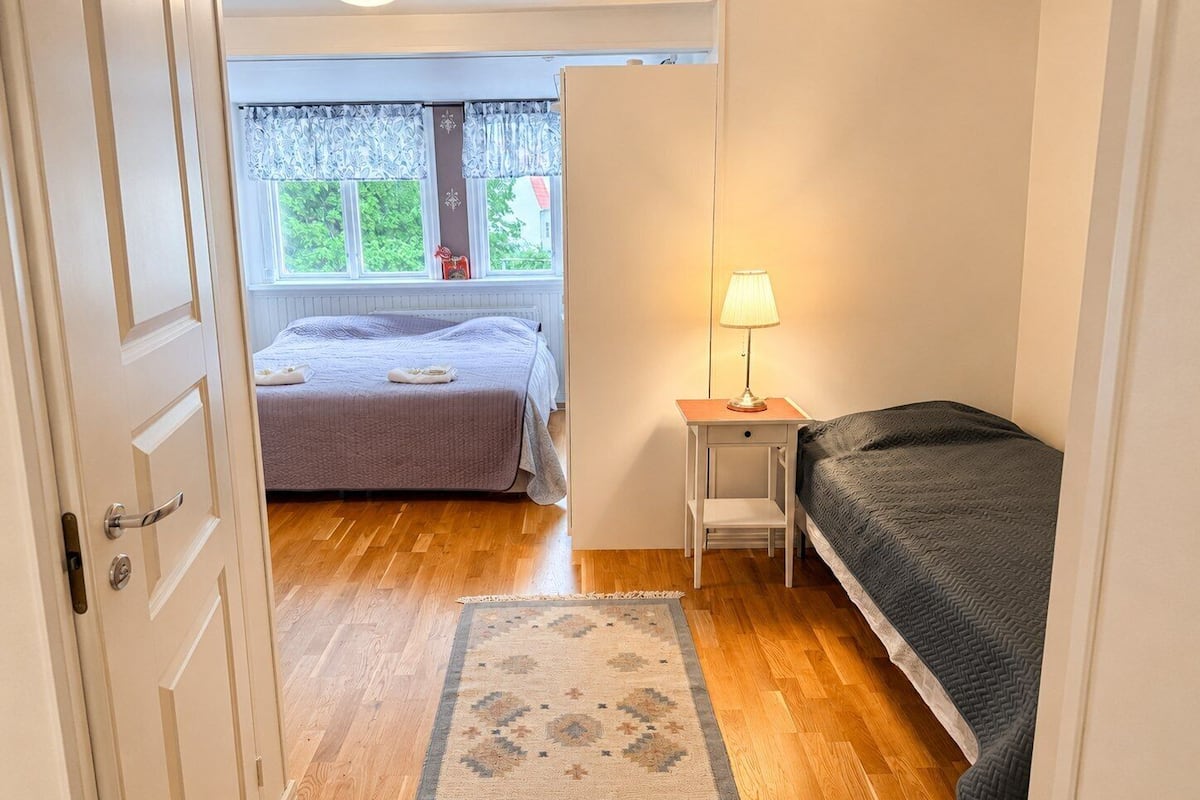 A bedroom with two beds, a window with curtains, a bedside table with a lamp, and a wooden floor.