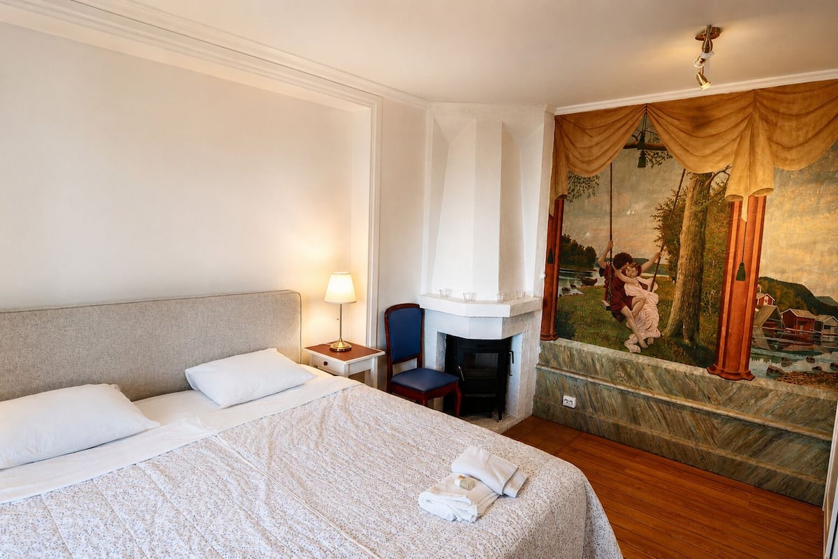 A hotel room with a bed, a nightstand, a lamp, a chair, a fireplace, and a mural.