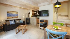 Ein modernes Wohnzimmer mit Sofa, Couchtisch und Esstisch. Es gibt eine Küchenzeile mit Herd, Spüle und Kühlschrank. Ein Fernseher ist an der Wand montiert und eine Treppe ist sichtbar.