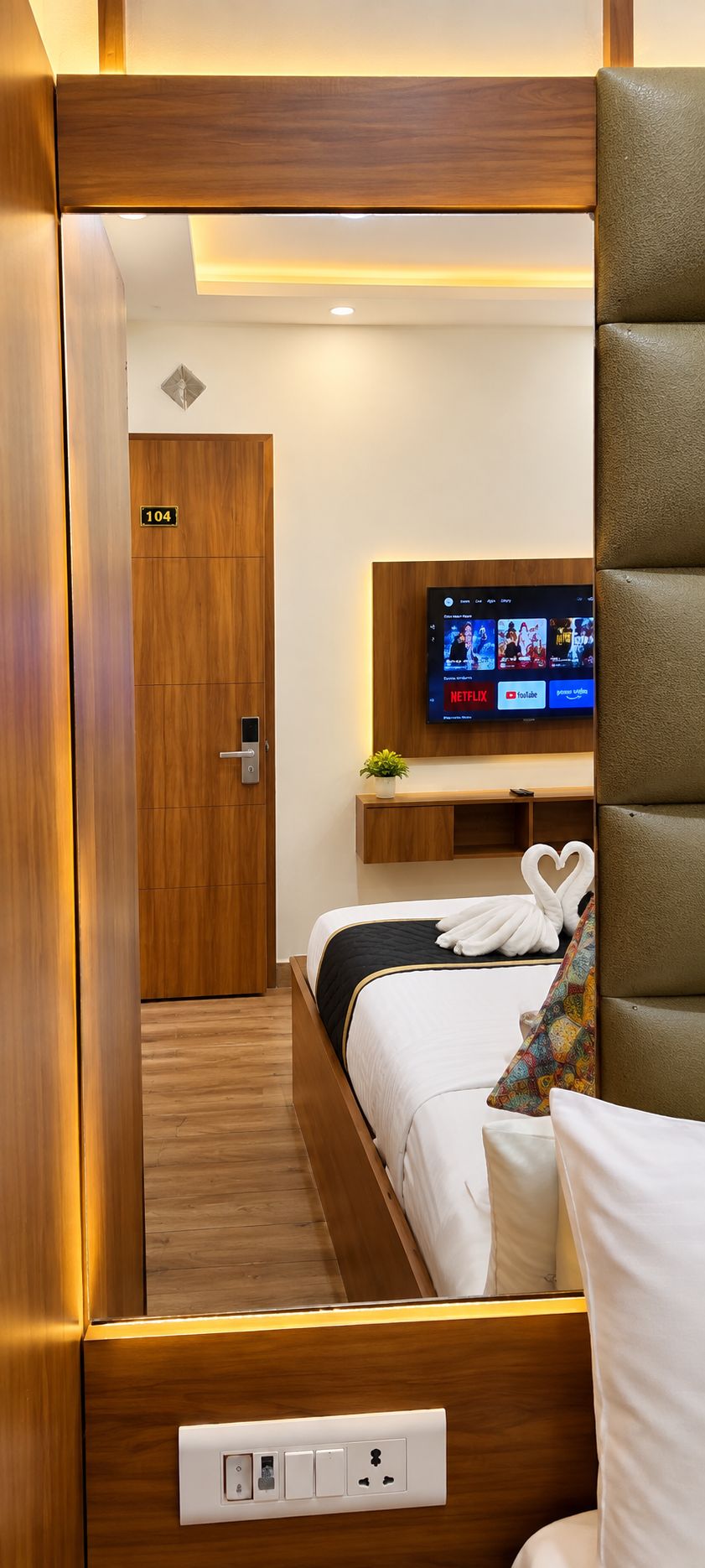 Habitación de hotel con cama, televisor montado en la pared, mesita de noche con planta y puerta con número.