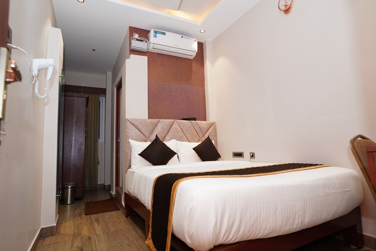 A hotel room with a bed, a wall-mounted air conditioner, a telephone, and a door leading to another room.