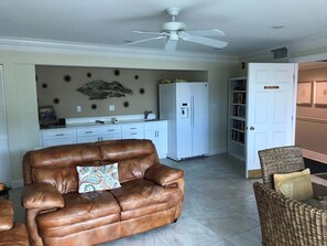 A living room with a brown leather sofa, a white refrigerator, a ceiling fan, and a bookshelf. - Waterfront 3BR condo with heated pool, wifi, and Marco Island views. (Marco Island)