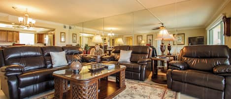 A spacious living room with leather sofas, a glass coffee table, and a large mirror reflecting the kitchen area.