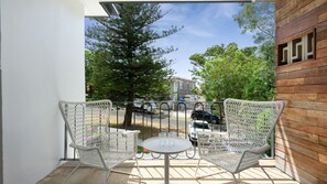 Ein Balkon mit zwei Korbstühlen und einem kleinen Tisch, mit Blick auf eine Straße mit parkenden Autos und Bäumen.