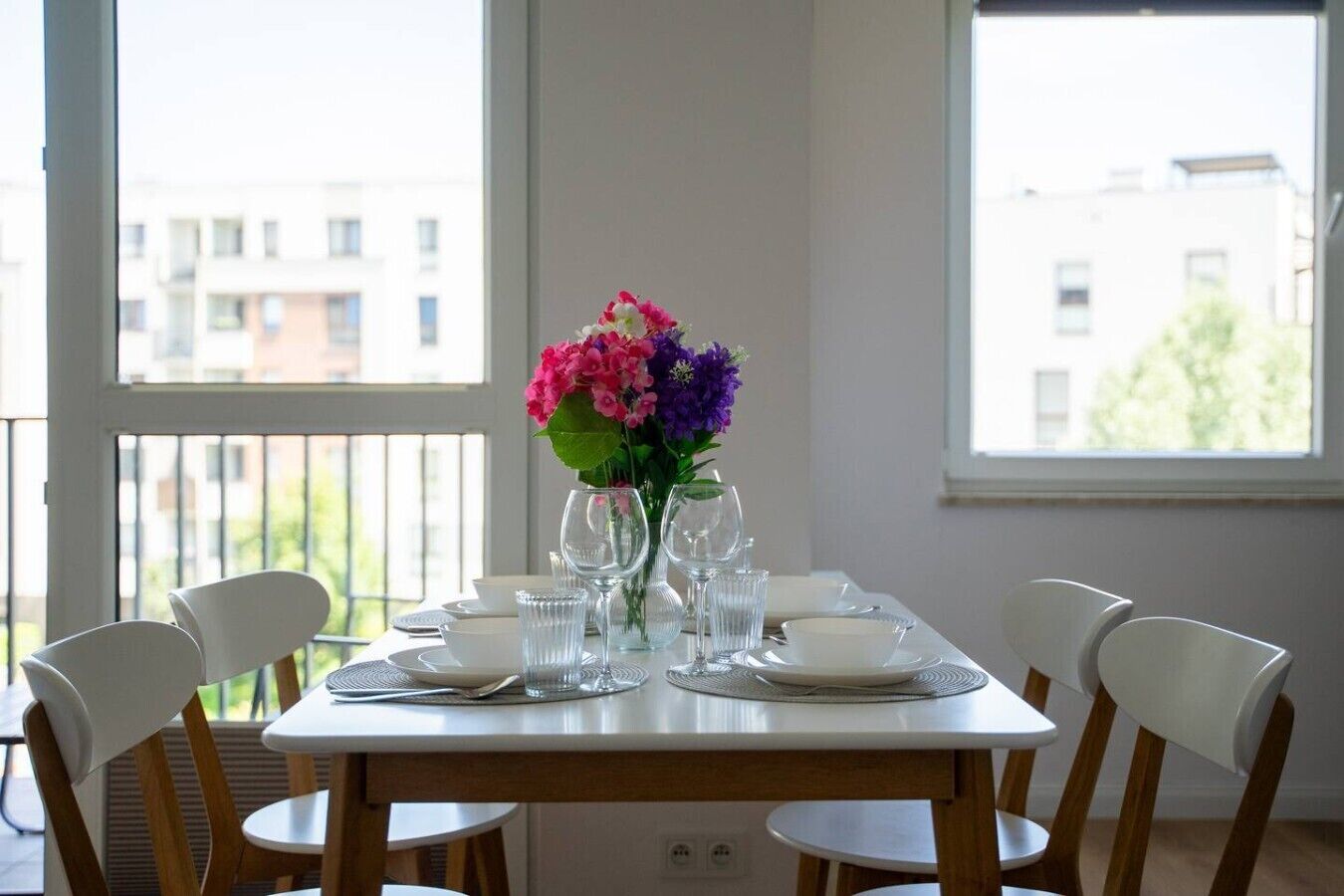 A dining table with chairs, a vase of flowers, and a view of buildings through large windows.